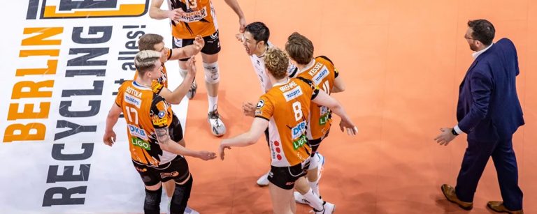 Mehrere Spieler der Berlin Recycling Heroes Volleyballmannschaft laufen kreisförmig auf einander zu in orangenen Trikots, rechts ist ein Trainer in einem blauen Anzug zu sehen