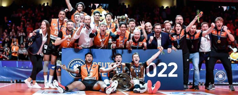 Gruppenfoto der Berlin Recycling Volleys zur Siegerehrung der deutschen Meisterschaft 2022, mit einem Transparent, mehreren Trainern sowie einer Trophäe in den Händen eines Spielers