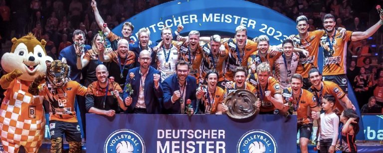 Gruppenfoto der Berlin Recycling Volleys zur Siegerehrung der deutschen Meisterschaft 2023, mit einem Transparent, Maskottchen, mehreren Trainern und Kindern und einer Trophäe in den Händen eines Spielers